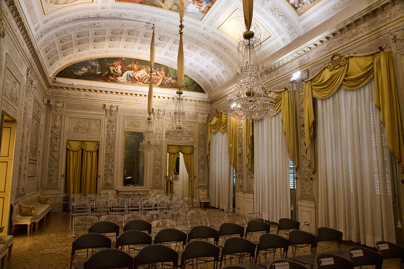 Il salone delle feste di Palazzo Milzetti. Si vedono stucchi, affreschi mitologici, finestre con pesanti tende ocra, e candelabri in cristallo.